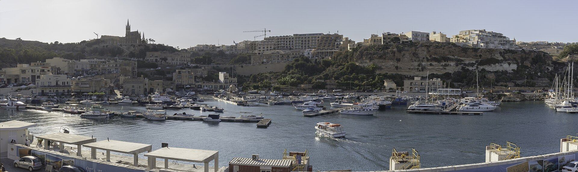 Panorama of Mġarr harbour on Gozo, boats at their moorings below the terraced hillside and the church of Our Lady of Lourdes above.