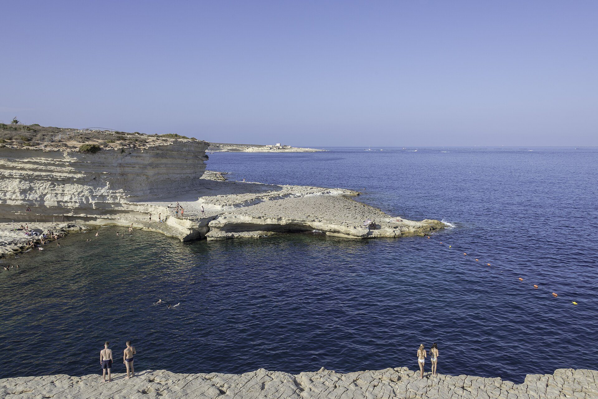 The flat pale limestone shelf at St Peter's Pool on the Delimara peninsula, with people standing at the water's edge
