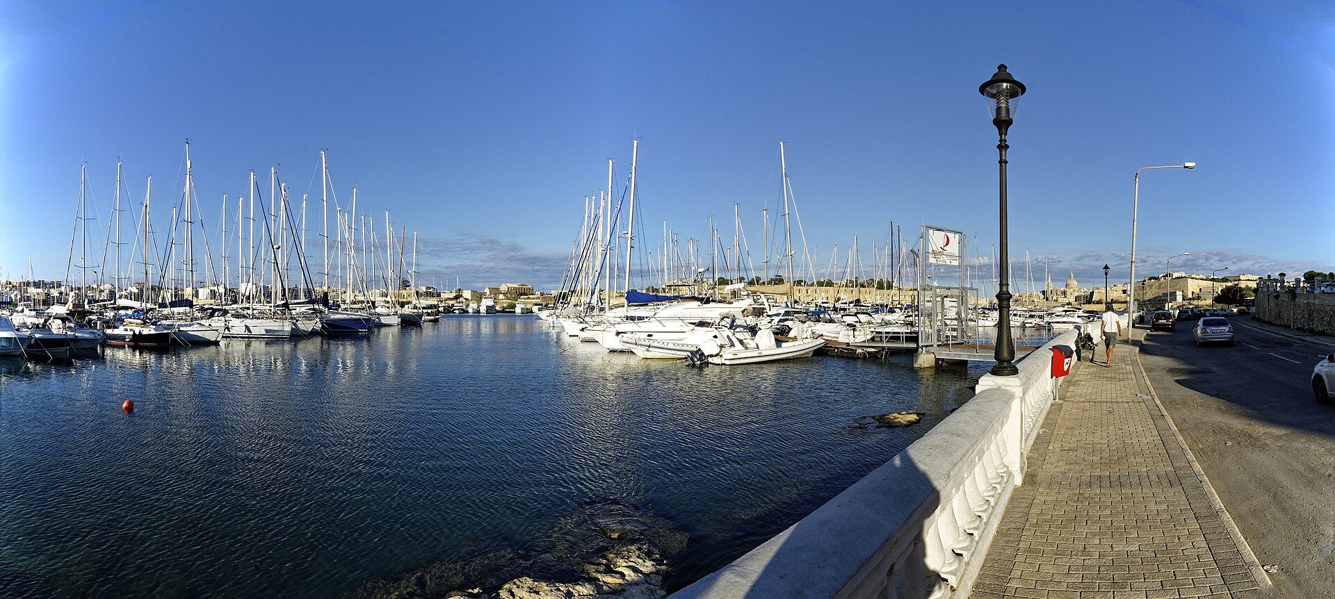 The Ta' Xbiex waterfront in evening light, ranks of sailing yacht masts along the quay and the Valletta bastions on the skyline beyond.